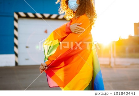 African American lesbian woman with medical face mask posing with lgbt pride flag. Curly woman during quarantine of coronavirus pandemic. African American lesbian woman with medical face mask posing with lgbt pride flag. Curly woman during quarantine of coronavirus pandemic. 101840441