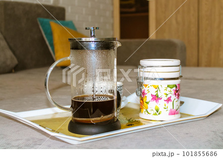 A glass coffee teapot and a ceramic sugar bowl stand on a tray on a gray sofa in the modern living room in a homely atmosphere. A cozy home at autumn season. Hot beverage to drink. Good morning. 101855686