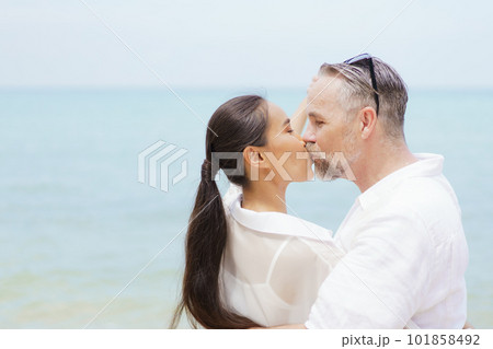 Happy Asian and caucasian white couple enjoy relaxing together at the beach during vacation or their honeymoon dating. Husband gently kissing his wife. 101858492