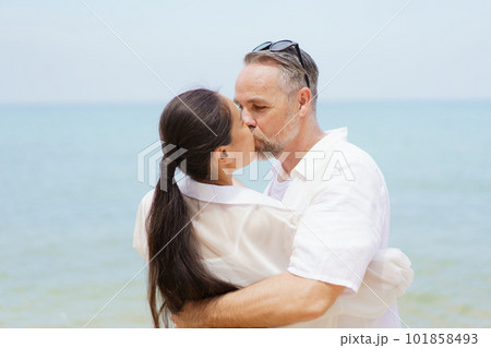 Happy Asian and caucasian white couple enjoy relaxing together at the beach during vacation or their honeymoon dating. Husband gently kissing his wife. 101858493