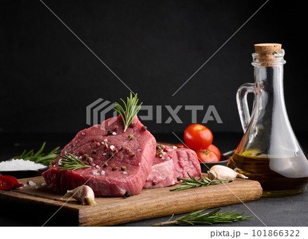 Raw piece of beef with spices pepper, rosemary sprig, salt and olive oil on a wooden board, black background Raw piece of beef with spices pepper, rosemary sprig, salt and olive oil on a wooden board, black background 101866322