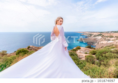 Blonde with long hair on a sunny seashore in a white flowing dress, rear view, silk fabric waving in the wind. Against the backdrop of the blue sky and mountains on the seashore. Blonde with long hair on a sunny seashore in a white flowing dress, rear view, silk fabric waving in the wind. Against the backdrop of the blue sky and mountains on the seashore. 101874097