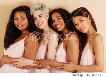 Group of smiling females with different skin tones against a beige background. Different women standing together in studio. Diverse cheerful females are hugging each other.	 101874819