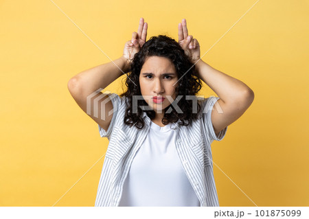 Portrait of strict serious woman with dark wavy hair showing bull horn gesture with fingers over head, looking hostile and threatening. Indoor studio shot isolated on yellow background. 101875099