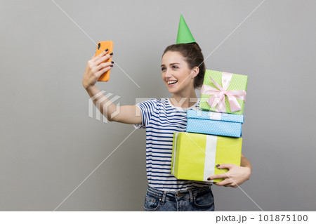 Portrait of friendly smiling woman wearing striped T-shirt and party cone, holding stack of present boxes and taking selfie, broadcasting livestream. Indoor studio shot isolated on gray background. Portrait of friendly smiling woman wearing striped T-shirt and party cone, holding stack of present boxes and taking selfie, broadcasting livestream. Indoor studio shot isolated on gray background. 101875100