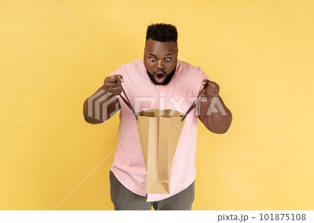 Portrait of man wearing pink shirt holding looking inside shopping bag, being astonished with cheap purchase or excellent gift. Indoor studio shot isolated on yellow background. 101875108
