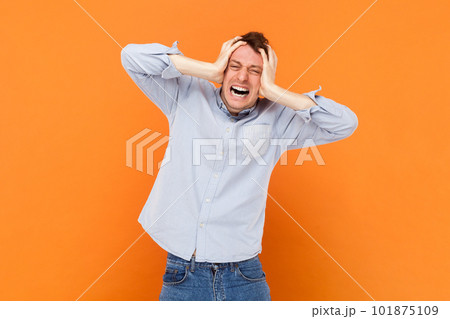 Portrait of upset sad young adult man standing with hands on head, suffering terrible headache, screaming from pain, wearing light blue shirt. Indoor studio shot isolated on orange background. 101875109