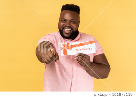 Portrait of excited amazed bearded man wearing pink shirt holding gift vaucher pointing to camera with toothy smile, you win present. Indoor studio shot isolated on yellow background. 101875110