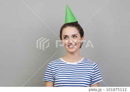 Portrait of delighted smiling dreamy woman wearing striped T-shirt and party cone looking away with satisfied smile, celebrating birthday. Indoor studio shot isolated on gray background. Portrait of delighted smiling dreamy woman wearing striped T-shirt and party cone looking away with satisfied smile, celebrating birthday. Indoor studio shot isolated on gray background. 101875112