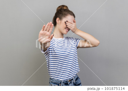 Portrait of afraid beautiful woman wearing striped T-shirt closing eyes with palm and showing stop hand gesture, turning face, does not want to see. Indoor studio shot isolated on gray background. 101875114
