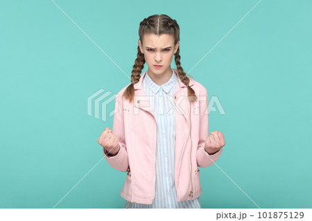 Portrait of angry aggressive attractive teenager girl with braids wearing pink jacket standing with clenched fits, arguing with somebody. Indoor studio shot isolated on green background. 101875129