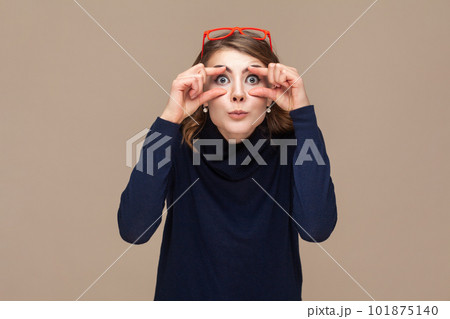 Portrait of funny surprised astonished woman with wavy hair in red red glasses on her head opens her eyes with fingers to see better. Indoor studio shot isolated on light brown background. 101875140