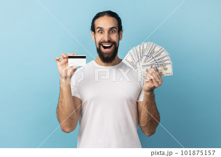 Amazed man wearing white T-shirt holding credit card and dollar banknotes, feeling rich with lot of money, excited to get bank loan for shopping. Indoor studio shot isolated on blue background. Amazed man wearing white T-shirt holding credit card and dollar banknotes, feeling rich with lot of money, excited to get bank loan for shopping. Indoor studio shot isolated on blue background. 101875157