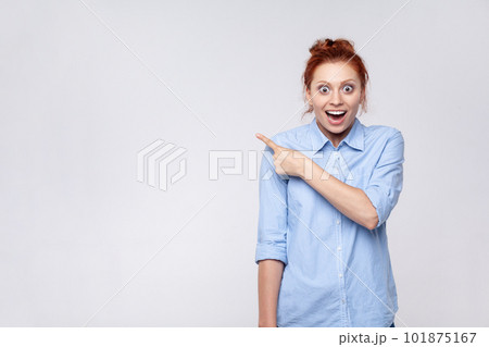 Portrait of shocked ginger woman wearing blue shirt looking at camera and pointing away, presenting copy space for advertisement or promotion. Indoor studio shot isolated on gray background. 101875167