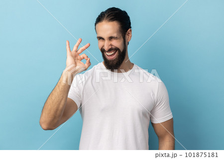 Portrait of satisfied happy man with beard wearing white T-shirt standing, looking at camera showing Ok sign gesture, expressing positive emotions. Indoor studio shot isolated on blue background. 101875181