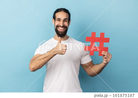 Portrait of happy man with beard wearing white T-shirt holding hashtag symbol, showing thumb up, recommending popular topics, internet trends. Indoor studio shot isolated on blue background. Portrait of happy man with beard wearing white T-shirt holding hashtag symbol, showing thumb up, recommending popular topics, internet trends. Indoor studio shot isolated on blue background. 101875187