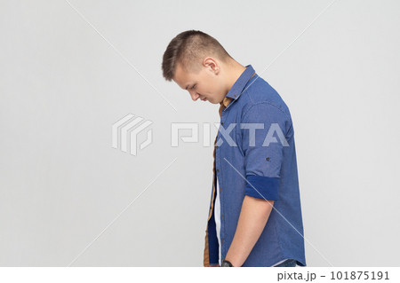 Side view portrait of upset teenager boy wearing blue shirt standing with his head down and looking, having with dissatisfied sadness face. Indoor studio shot isolated on gray background. Side view portrait of upset teenager boy wearing blue shirt standing with his head down and looking, having with dissatisfied sadness face. Indoor studio shot isolated on gray background. 101875191