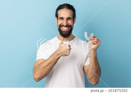 Portrait of man with beard wearing white T-shirt holding dental aligner retainer, dental clinic for beautiful teeth, showing thumb up. Indoor studio shot isolated on blue background. Portrait of man with beard wearing white T-shirt holding dental aligner retainer, dental clinic for beautiful teeth, showing thumb up. Indoor studio shot isolated on blue background. 101875202