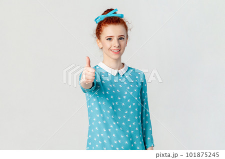 Portrait of satisfied positive optimistic red haired woman with bun hairstyle, standing showing like gesture, recommend, wearing blue dress. Indoor studio shot isolated on gray background. Portrait of satisfied positive optimistic red haired woman with bun hairstyle, standing showing like gesture, recommend, wearing blue dress. Indoor studio shot isolated on gray background. 101875245