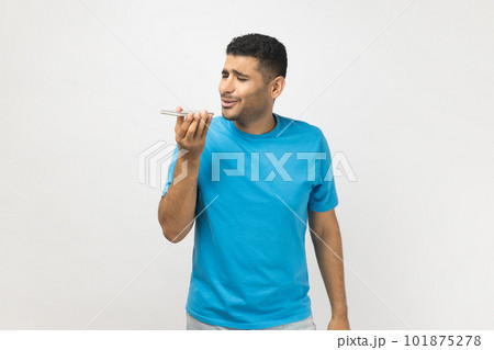 Portrait of handsome unshaven man wearing blue T- shirt standing holding smart phone, recording voice message, or using voice assistant. Indoor studio shot isolated on gray background. 101875278