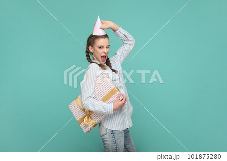 Portrait of excited cheerful teenager girl with braids wearing striped shirt and party cone, holding present box, gets gift on her birthday. Indoor studio shot isolated on green background. 101875280