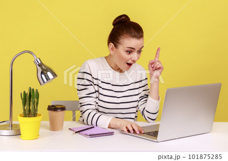 Amazed woman manager in striped shirt looking surprised by genius idea, raising finger up inspired while working on laptop. Indoor studio studio shot isolated on yellow background. 101875285