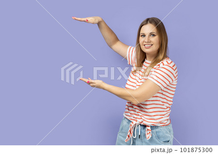 Portrait of satisfied smiling blond woman wearing striped T-shirt presenting area between hands for advertisement, showing huge size. Indoor studio shot isolated on purple background. Portrait of satisfied smiling blond woman wearing striped T-shirt presenting area between hands for advertisement, showing huge size. Indoor studio shot isolated on purple background. 101875300