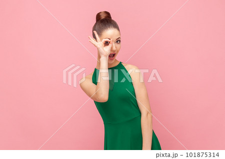 Portrait of funny childish young adult woman standing covering her eye with ok gesture, looking at camera, showing okay sign, wearing green dress. Indoor studio shot isolated on pink background. 101875314