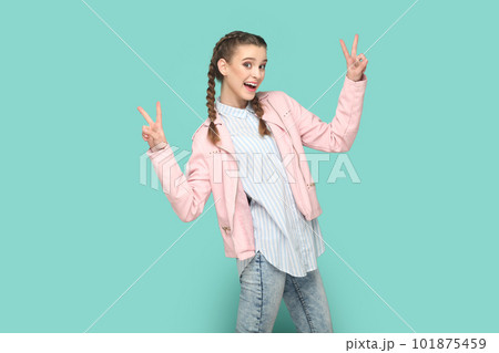 Portrait of excited optimistic cheerful teenager girl with braids wearing pink jacket showing v signs, being happy, being in high spirit. Indoor studio shot isolated on green background. Portrait of excited optimistic cheerful teenager girl with braids wearing pink jacket showing v signs, being happy, being in high spirit. Indoor studio shot isolated on green background. 101875459