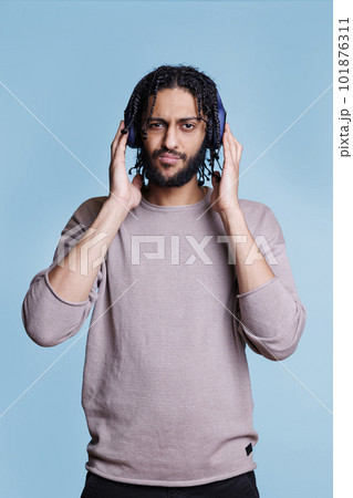 Cool arab man listening to music while touching headphones on head and looking at camera. Confident person with serious face expression enjoying music in earphone portrait 101876311