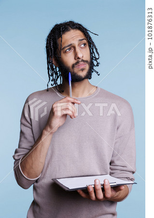 Puzzled arab man thinking while checking plan on clipboard and looking away. Pensive young adult person in doubt reading checklist and holding pen while standing in thoughtful pose 101876313