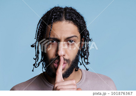 Arab man making silent gesture with serious facial expression and looking at camera. Young person with raised eyebrow showing quiet sign while holding finger on lips portrait 101876367