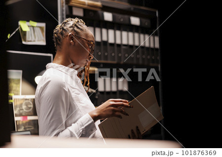 Thoughtful african american woman detective reading criminal investigation documentation in agency office room. Law enforcement professional studying criminologist report at night Thoughtful african american woman detective reading criminal investigation documentation in agency office room. Law enforcement professional studying criminologist report at night 101876369