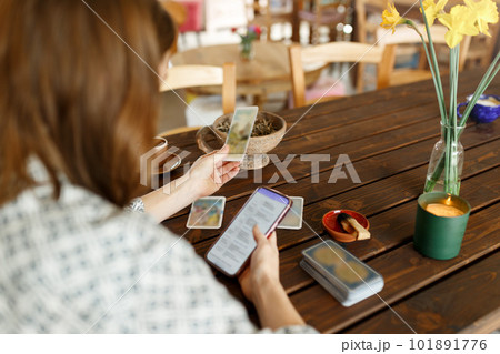 Beautiful young woman is guessing on cards with tarot, runes on wooden table and uses an online app in phone to interpret divination, home interior,candles and dry herbs.. 101891776