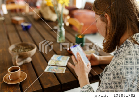 Beautiful young woman is guessing on cards with tarot, runes on wooden table and uses an online app in phone to interpret divination, home interior,candles and dry herbs.. 101891782