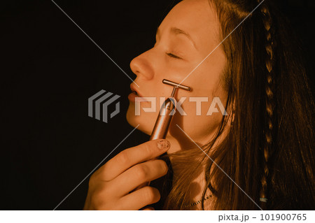 Young beautiful white woman, girl is using vibrating golden facial massager on dark black background with copy space. Preservation prolongation of natural beauty youth Female skin care with pleasure. 101900765