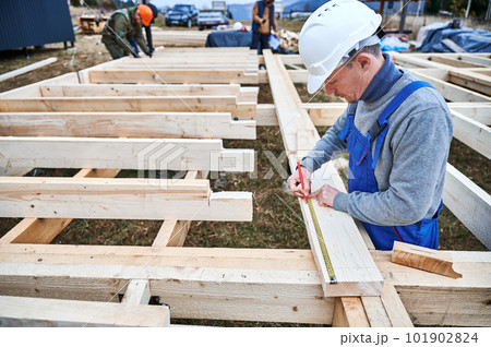 Man worker building wooden frame house on pile foundation. Carpenter using tape measure for measuring wooden planks and making marks with pencil. Carpentry concept. Man worker building wooden frame house on pile foundation. Carpenter using tape measure for measuring wooden planks and making marks with pencil. Carpentry concept. 101902824