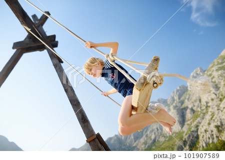 Little boy having fun on large swing on the shore in the Boka Kotor Bay of the Adriatic Sea in the Balkan Mountains, Montenegro. Happy child on a sea resort on sunny summer holidays day. Little boy having fun on large swing on the shore in the Boka Kotor Bay of the Adriatic Sea in the Balkan Mountains, Montenegro. Happy child on a sea resort on sunny summer holidays day. 101907589