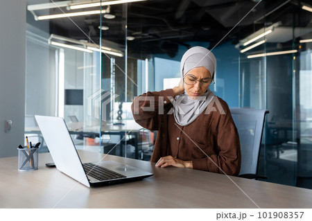 Tired young arab businesswoman in hijab working on project overtime in office at laptop and holding neck, grimacing in pain, massaging with hands. 101908357