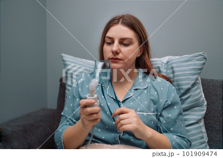 Young sick woman putting on nebulizer mask on face to make inhalation, using an inhaler at home. Medical inhaler emergency equipment, asthmatic attack 101909474