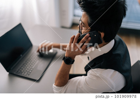 High-angle rear view of Indian bearded business man in glasses working at laptop sitting at desk and talking on smartphone by window. Handsome businessman in elegant formalwear talking to client High-angle rear view of Indian bearded business man in glasses working at laptop sitting at desk and talking on smartphone by window. Handsome businessman in elegant formalwear talking to client 101909978