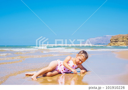 Cute little girl in pink swimsuit with strawberries lying in sand and play with water at the seashore against blue sky Cute little girl in pink swimsuit with strawberries lying in sand and play with water at the seashore against blue sky 101910226