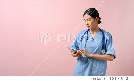 Beautiful female doctor writing on tablet while standing on pink isolated background. 101919385