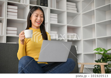 Adorable young woman using laptop and drinking coffee while sitting on the sofa Adorable young woman using laptop and drinking coffee while sitting on the sofa 101920513