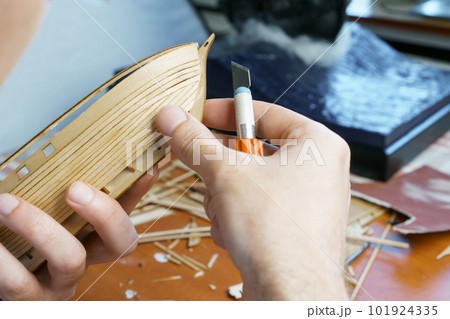 Hands man cutting out details for ship model with clerical knife from plywood. Process of building toy ship, handicraft. 101924335
