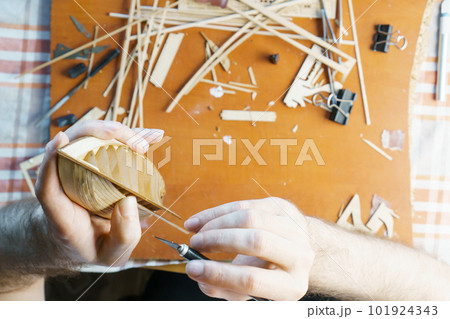 Hands of man removing excess glue from plywood details for ship model with utility knife. Process of building toy ship 101924343
