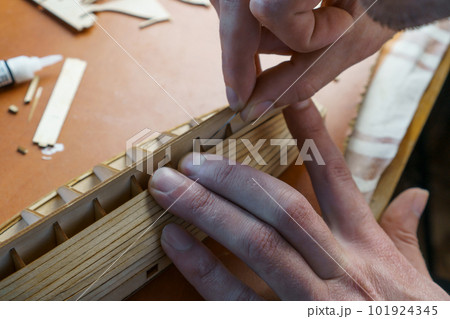 Hands of man gluing plywood details for ship model with glue, holding with fingers. Process of building toy ship 101924345