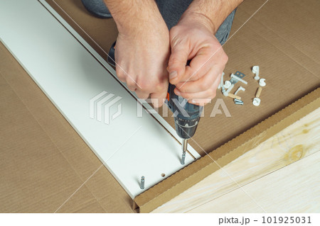 Male hands hold cordless electric screwdriver, screw to fix walls of white drawer, assembling dresser, wardrobe on floor 101925031