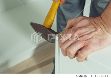 Male hands hold metal hammer with yellow plastic handle, driving dowel, assembling white drawer of dresser or wardrobe Male hands hold metal hammer with yellow plastic handle, driving dowel, assembling white drawer of dresser or wardrobe 101925041