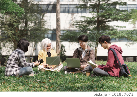 A multiethnic group of asian university students is seen studying together while sitting on the grass near the campus. 101937073